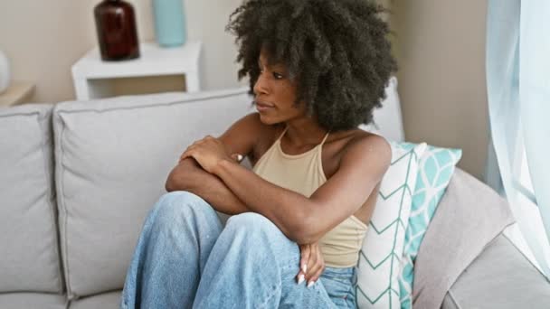 Африканская американка сидит на диване с грустным выражением лица и скрещенными руками дома - Кадры, видео