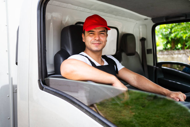Boldog profi kamionsofőr asszisztensével piros sapkát visel, mosolyog, belenéz a kamerába a teherautó ablakából, mielőtt kiszállítja a csomagot. A teherautó-vezető és a szállítási szolgáltatás koncepciója. - Fotó, kép