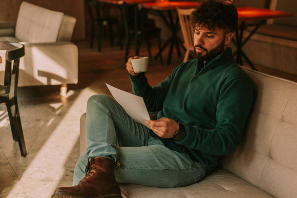 Νεαρός όμορφος αρχηγός ομάδας επιχειρήσεων διαβάζει τα αποτελέσματα της ομαδικής δουλειάς ενώ πίνει τον πρώτο του καφέ νωρίς το πρωί στο καφέ μπαρ - Φωτογραφία, εικόνα