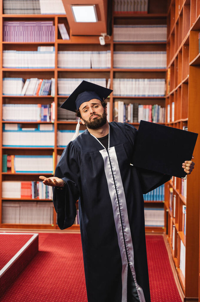 Joven graduado vistiendo su gorra y vestido mirando triste mientras sostiene su título universitario - Foto, imagen