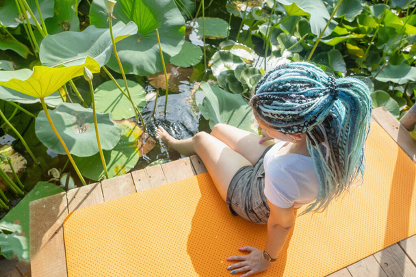 jeune belle femme avec des serrures afro bleues reposant sur un tapis de yoga sur un perce-bois sur un lac de lotus profitant de la nature, jambes dans l'eau - Photo, image