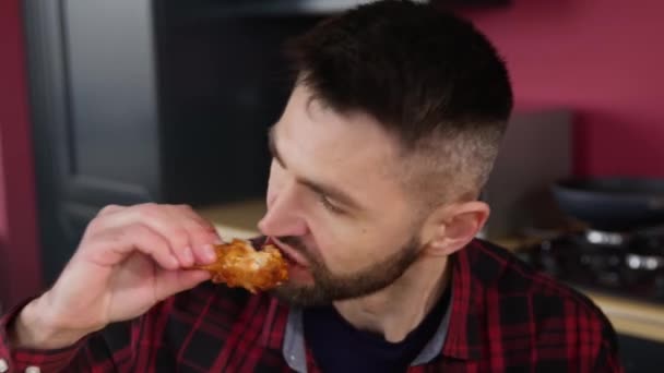 Közelről. Éhes fiatal barna férfi kockás ingben csirkeszárnyakat eszik a bal és a jobb kezéről, miközben élvezi. A gyorskaja elfogyasztásának fogalma. - Felvétel, videó