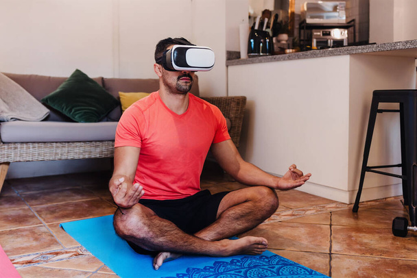 Hombre latino meditando usando auriculares de realidad virtual en casa en México América Latina. Personas hispanas positivas en apartamentos que practican yoga con tecnología moderna VR - Foto, Imagen