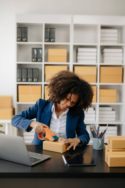 Startup small business SME. Entrepreneur African woman using laptop to receive and check online orders. Young female small business owner packing parcels to ship them to customers - Φωτογραφία, εικόνα