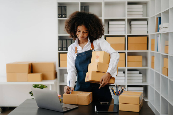 Startup small business SME. Entrepreneur African woman receiving and checking online orders. Young female small business owner packing parcels to ship them to customers - Valokuva, kuva