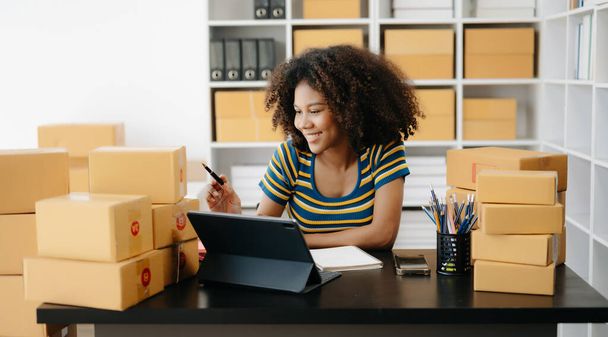 Startup small business SME. Entrepreneur African woman receiving and checking online orders. Young female small business owner packing parcels to ship them to customers - Фото, изображение