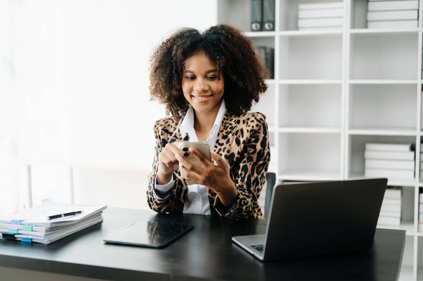 Young African woman with afro hair, wearing leopard jacket while working on office and using mobile  - Fotografie, Obrázek