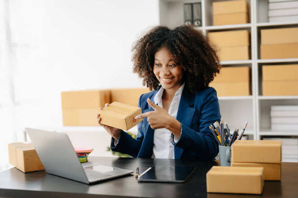 Startup small business SME. Entrepreneur African woman using laptop to receive and check online orders. Young female small business owner packing parcels to ship them to customers - Φωτογραφία, εικόνα