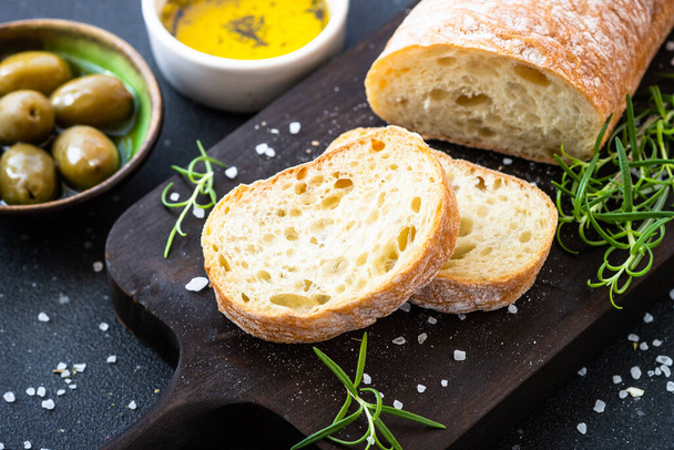 Ciabatta bread on wooden board with olive oil, olives and herbs on black. Mediterranean food. - Photo, Image