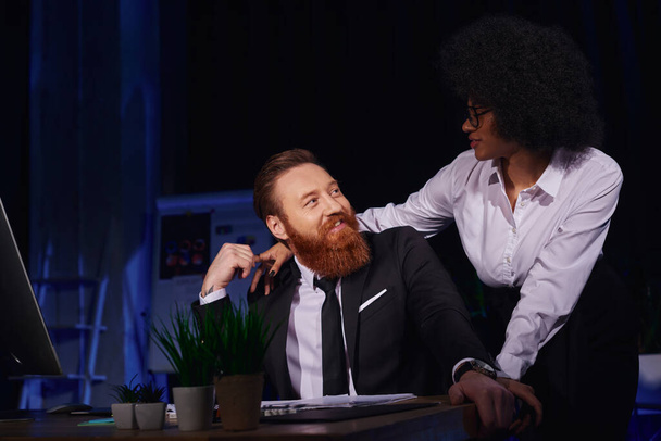 african american woman embracing happy bearded businessman in night office, love affair at work - Foto, afbeelding