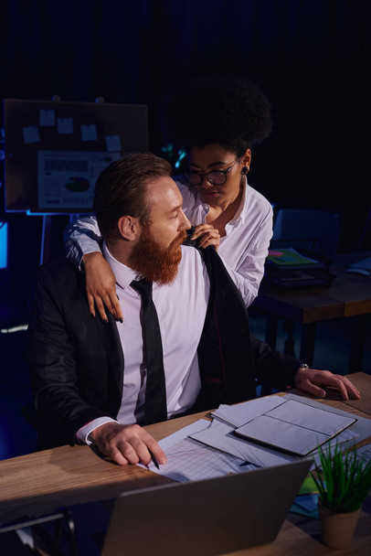 passionate african american woman undressing bearded businessman near documents and laptop in office - Photo, Image