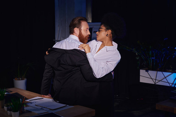pareja interracial apasionada en el desgaste formal abrazo en el lugar de trabajo en la oficina por la noche, romance de trabajo - Foto, imagen