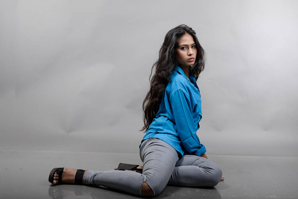 Modello giovane donna indiana in abbigliamento casual su sfondo grigio - foto stock. Modello lungo per capelli neri con camicia blu, jeans grigi e tacchi neri. - Foto, immagini