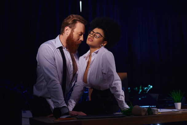 mujer afroamericana apasionada en anteojos seduciendo a hombre de negocios barbudo en la oficina por la noche - Foto, Imagen