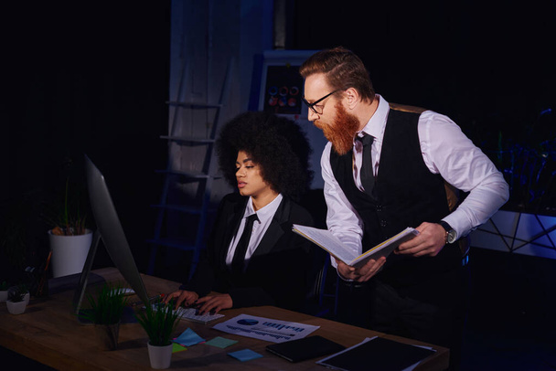 african american woman typing on computer near bearded businessman with notebook in office at night - 写真・画像