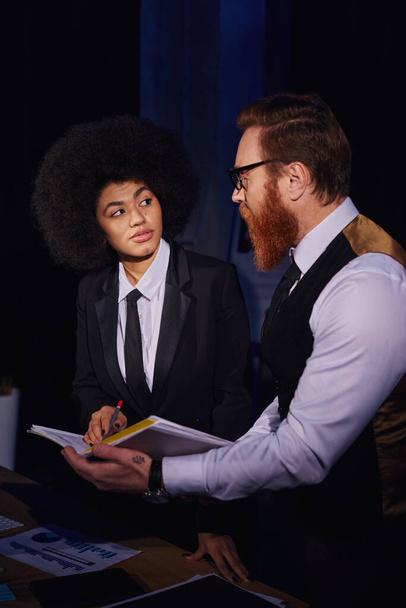 interracial entrepreneurs looking at each other while working with documents at night in office - Photo, image