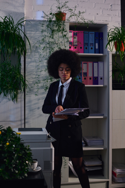donna afroamericana in abito formale in piedi con documenti cartacei vicino fotocopiatrice di notte in ufficio - Foto, immagini