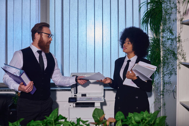 bärtiger Geschäftsmann mit Brille übergibt Dokumenten an junge afrikanisch-amerikanische Sekretärin in der Nähe eines Kopierers - Foto, Bild