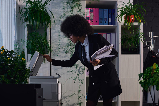 κομψή Αφροαμερικανίδα επιχειρηματίας με έγγραφα που ανοίγουν φωτοτυπικό μηχάνημα τη νύχτα στο γραφείο - Φωτογραφία, εικόνα