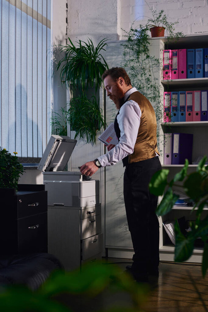 vista laterale di barbuto elegante uomo d'affari che lavora con fotocopiatrice in ufficio in serata - Foto, immagini