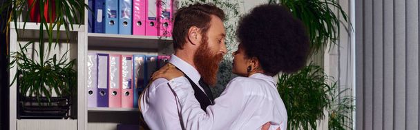 pareja interracial abrazando en la noche cerca de rack con carpetas en la noche, el amor en el trabajo, pancarta - Foto, Imagen