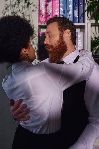 bearded businessman hugging young african american secretary, night romance in office - 写真・画像