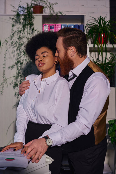 hombre de negocios abrazando apasionada mujer afroamericana con los ojos cerrados cerca de fotocopiadora en la oficina de noche - Foto, Imagen