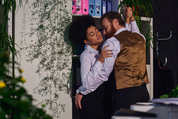 pareja interracial apasionada abrazando cerca de plantas decorativas en la oficina por la noche, romance de trabajo - Foto, imagen