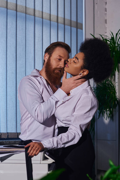 Ofisteki fotokopi makinesinin yanında seksi Afrikalı Amerikalı bir kadının boynuna sarılan sakallı ve dövmeli bir işadamı. - Fotoğraf, Görsel