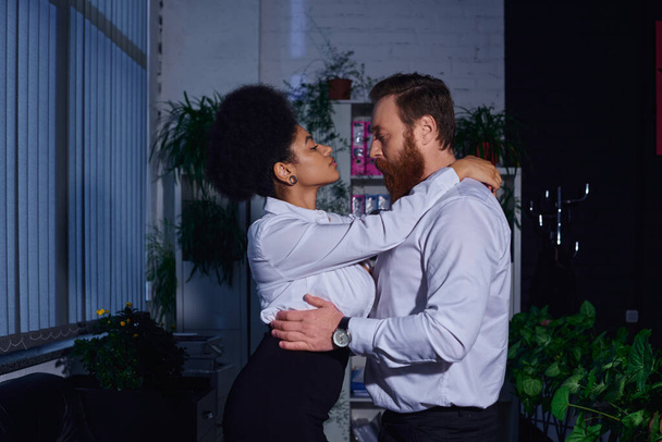 vista lateral de la encantadora secretaria afroamericana seduciendo a hombre de negocios barbudo en la oficina por la noche - Foto, Imagen