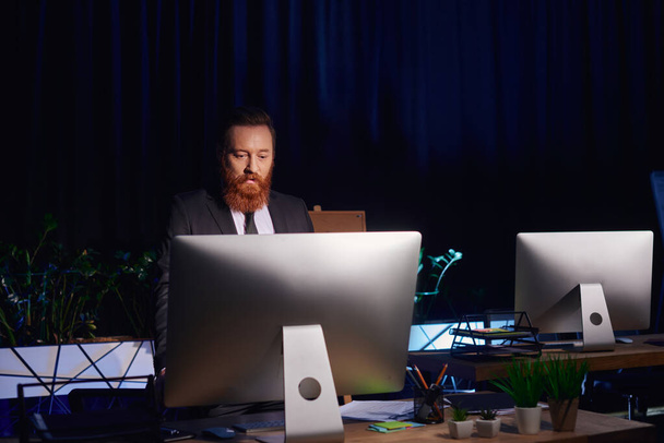 hombre de negocios barbudo serio mirando el monitor de la computadora mientras trabaja hasta tarde en la oficina, turno de noche - Foto, imagen