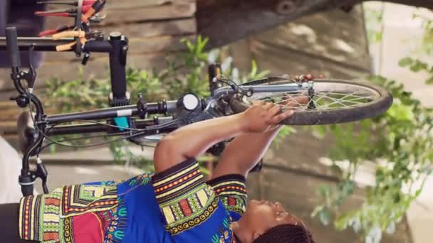 Verticale video Actieve Afrikaans-Amerikaanse vrouw voorzichtig losmaken en verwijderen van beschadigde fietswiel. Sportliefhebbende vrouwelijke fietser zorgvuldig ontmantelen band van fiets voorvork voor onderhoud in - Video