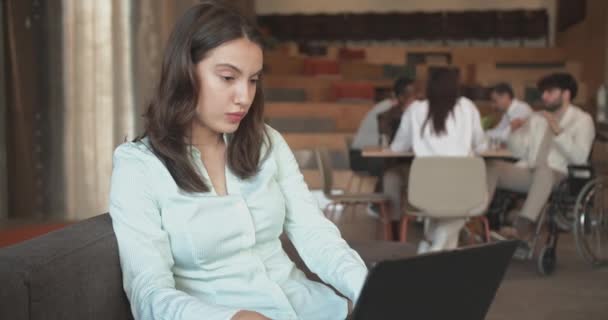 Όμορφη νεαρή γυναίκα που εργάζεται για το laptop της, ενώ στηρίζεται στον καναπέ, οι συνάδελφοί της συζητούν επιχειρηματικές ιδέες στο καφέ μπαρ στο γραφείο, φορτηγά - Πλάνα, βίντεο