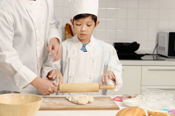 Mutlu Asyalı oğul ve aşçı üniformalı baba, mutfakta birlikte yemek pişiren şapkalı yakışıklı çocuk merdaneyle hamuru yoğuruyor, aile içinde fırın yapan ebeveyn ve çocuk. Çocuk öğrenci ekmek pişirmeyi öğreniyor.. - Fotoğraf, Görsel