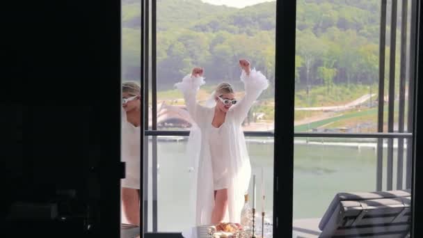 Una sposa felice in occhiali da sole bianchi, una camicia da notte bianca e un accappatoio di seta bianca sta ballando sul balcone. La mattina della sposa felice. - Filmati, video