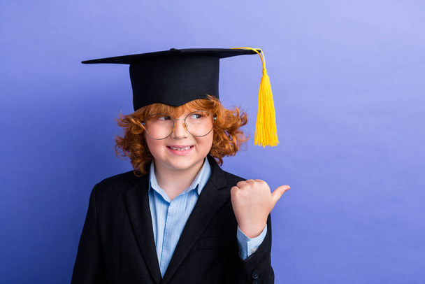 Portrait of small intelligent boy beaming smile look indicate finger empty space wear mortarboard hat isolated on purple color background. - Foto, imagen