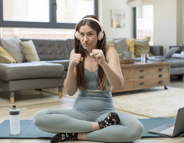 young adult woman practicing yoga looking confident, angry, strong and aggressive, with fists ready to fight in boxing position - Фото, зображення