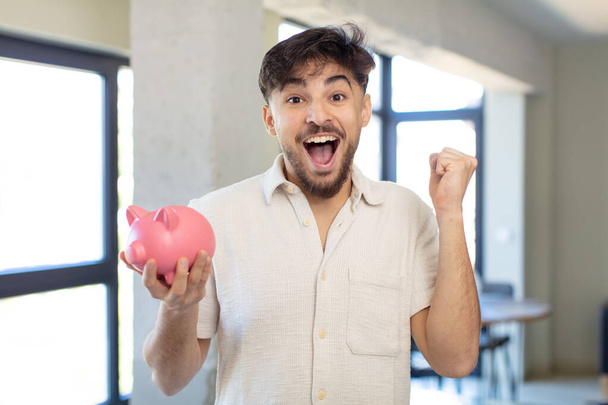 young handsome man feeling shocked,laughing and celebrating success. piggy bank concept - 写真・画像