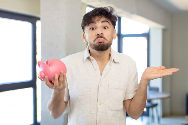 young handsome man shrugging, feeling confused and uncertain. piggy bank concept - Foto, afbeelding