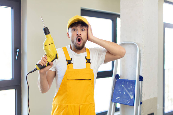 young handsome man feeling extremely shocked and surprised. handyman and drill concept - Valokuva, kuva