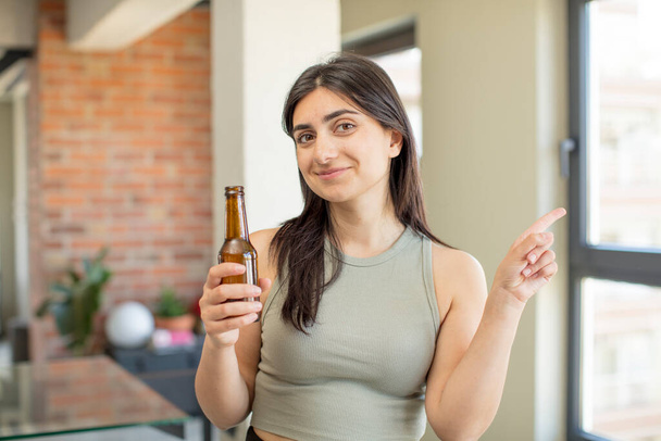 young woman smiling cheerfully, feeling happy and pointing to the side. beer bottle - Zdjęcie, obraz