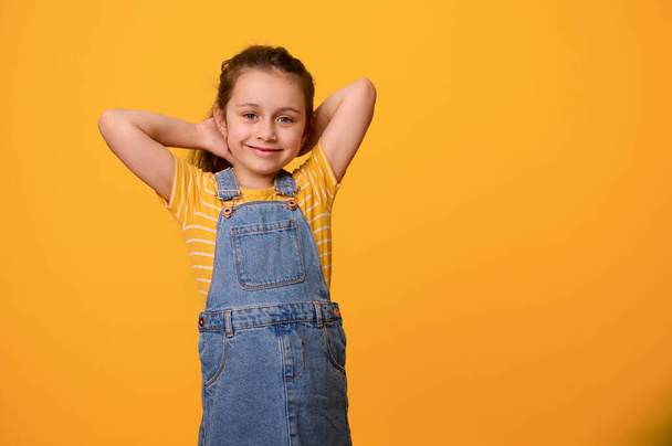 Красива харизматична весела емоційна дівчинка 6 років, одягнена в блакитний джинсовий комбінезон, посміхається дивлячись на камеру, ізольована на помаранчевому студійному фоні. Люди. Поняття. Вирази обличчя - Фото, зображення