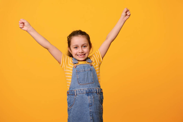 Портрет счастливой улыбающейся веселой харизматичной маленькой дошкольницы, одетой в синее джинсовое платье, сжимающей кулаки и поднимающей руки, выражающей позитивные эмоции, изолированной на фоне оранжевой студии - Фото, изображение