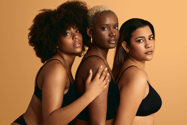 Side view of young multiracial women wearing black lingerie standing close in row and looking at camera against beige background - 写真・画像