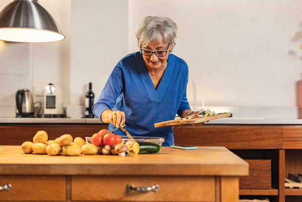 jolie femme caucasienne senior préparant un délicieux repas dans sa cuisine - femme retraitée aux cheveux gris jouissant de temps libre - - Photo, image