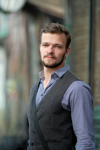 Street portrait of a young handsome man with a small beard and mustache in a vest on a warm summer day - Photo, Image