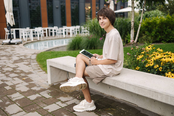 Teenage boy using tablet outdoors. Happy student have rest during the break in park near the campus. High quality photo - Photo, image