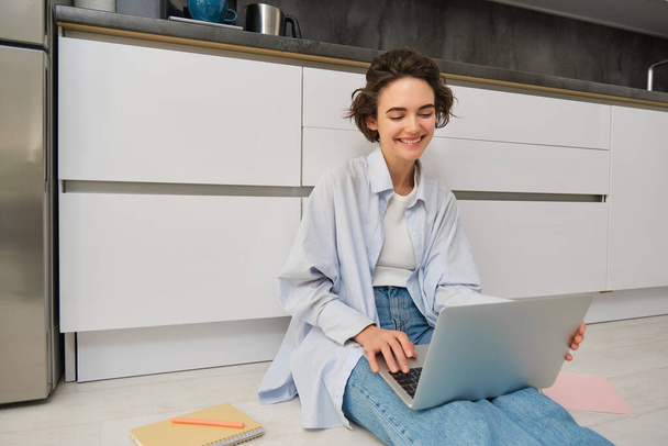 Πορτρέτο της νεαρής γυναίκας που χρησιμοποιεί φορητό υπολογιστή, κάθεται στο πάτωμα της κουζίνας, κάνει την εργασία της, σε απευθείας σύνδεση συνομιλία με τους φίλους στον υπολογιστή. - Φωτογραφία, εικόνα