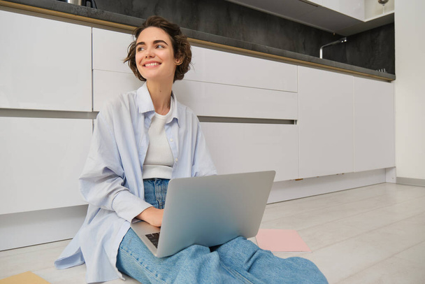 Mujer joven trabaja desde casa en el suelo de la cocina, hace los deberes en el ordenador portátil, el uso de Internet en la computadora, freelancing. - Foto, Imagen