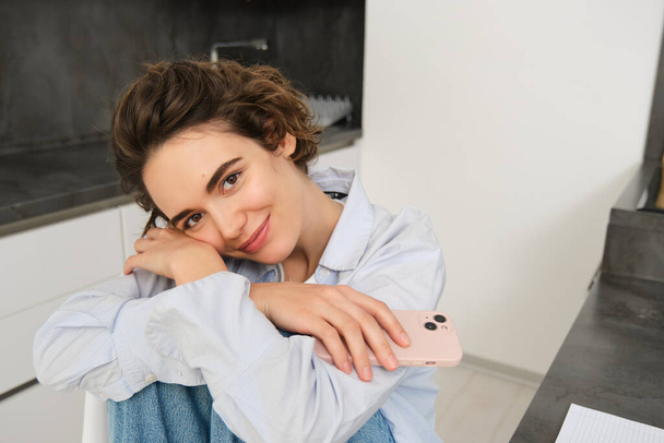 Feminine smiling girl sitting on kitchen chair with smartphone, daydreaming, looking happy. Mobile connection and people concept - Photo, Image
